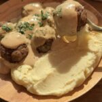 A wooden plate featuring juicy homemade meatballs covered in creamy gravy, served alongside buttery mashed potatoes, garnished with freshly chopped parsley for a flavorful and visually appealing dish.