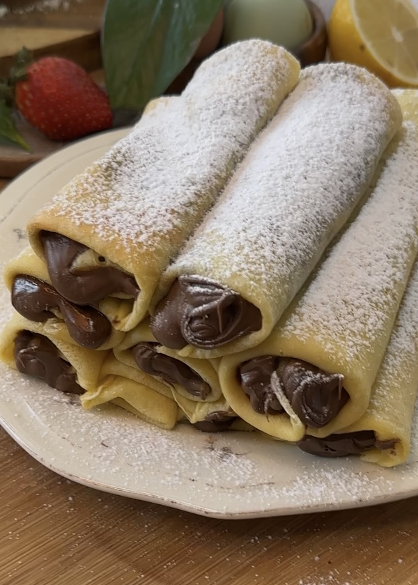 Stack of Nutella crepe rolls on a plate with Nutella oozing out and powdered sugar on top, ready to serve