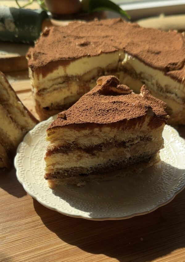 A slice of tiramisu cheesecake on a plate with biscuit crust, coffee-soaked ladyfingers, creamy no-bake cheesecake layers, and a dusting of cocoa powder.