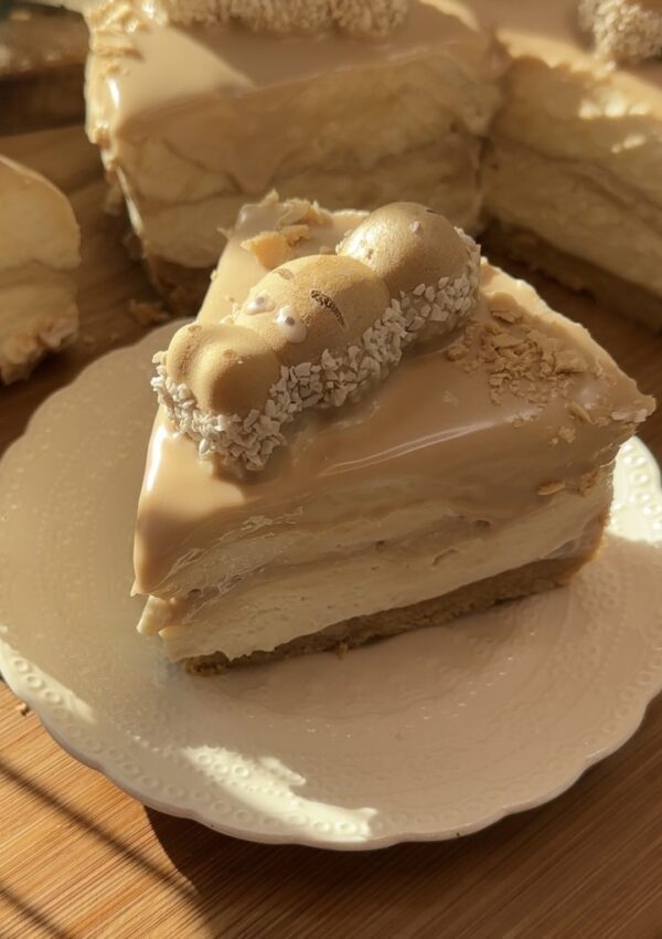 A slice of Happy Hippo No-Bake Cheesecake on a plate, featuring a crunchy biscuit base, creamy hazelnut cheesecake filling, Kinder-filled interior, and topped with Kinder Bueno spread, wafer rolls, Happy Hippo, and pearl sugar